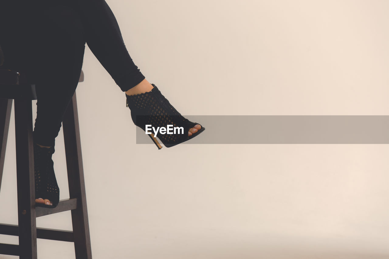 Low section of woman wearing high heels while sitting on stool against wall