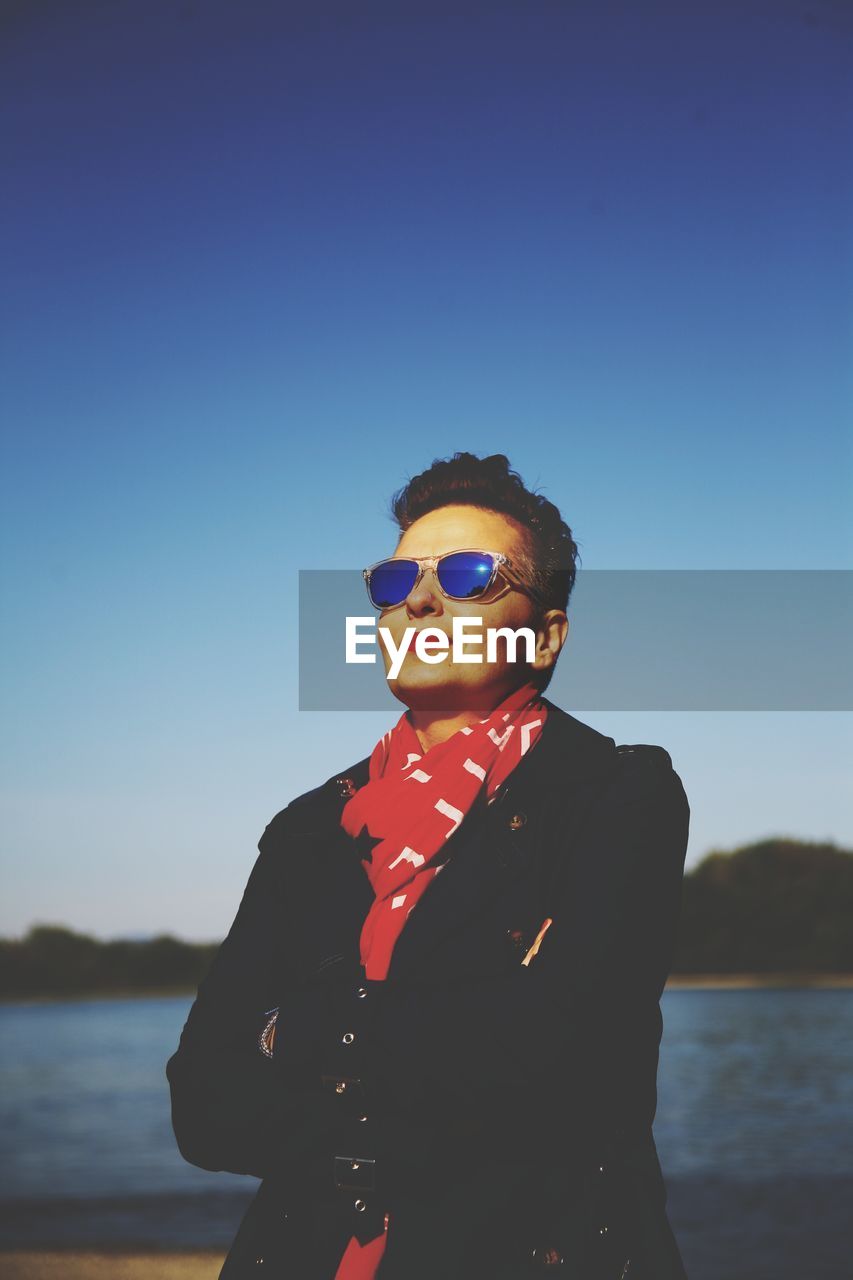 Smiling woman wearing sunglasses while standing by lake against clear blue sky