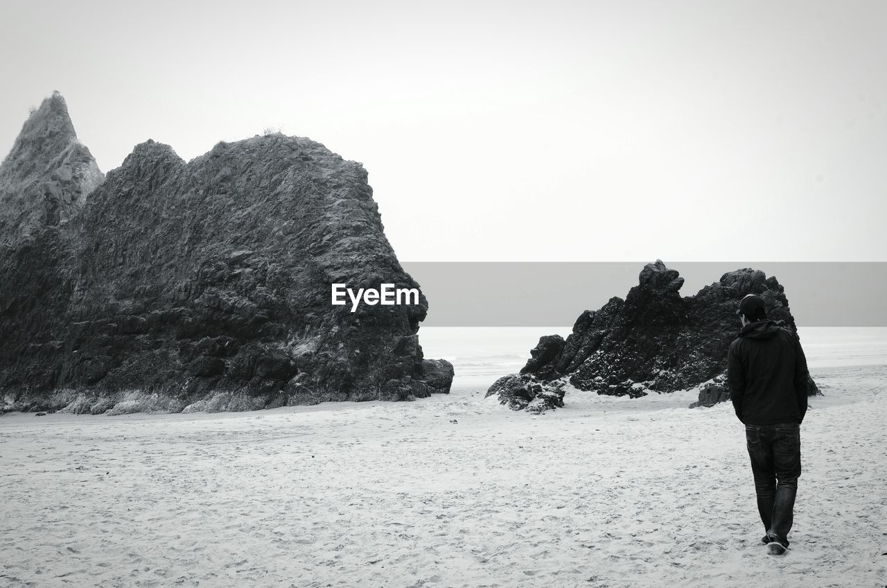 Rear view of man walking towards rock formations at beach against clear sky