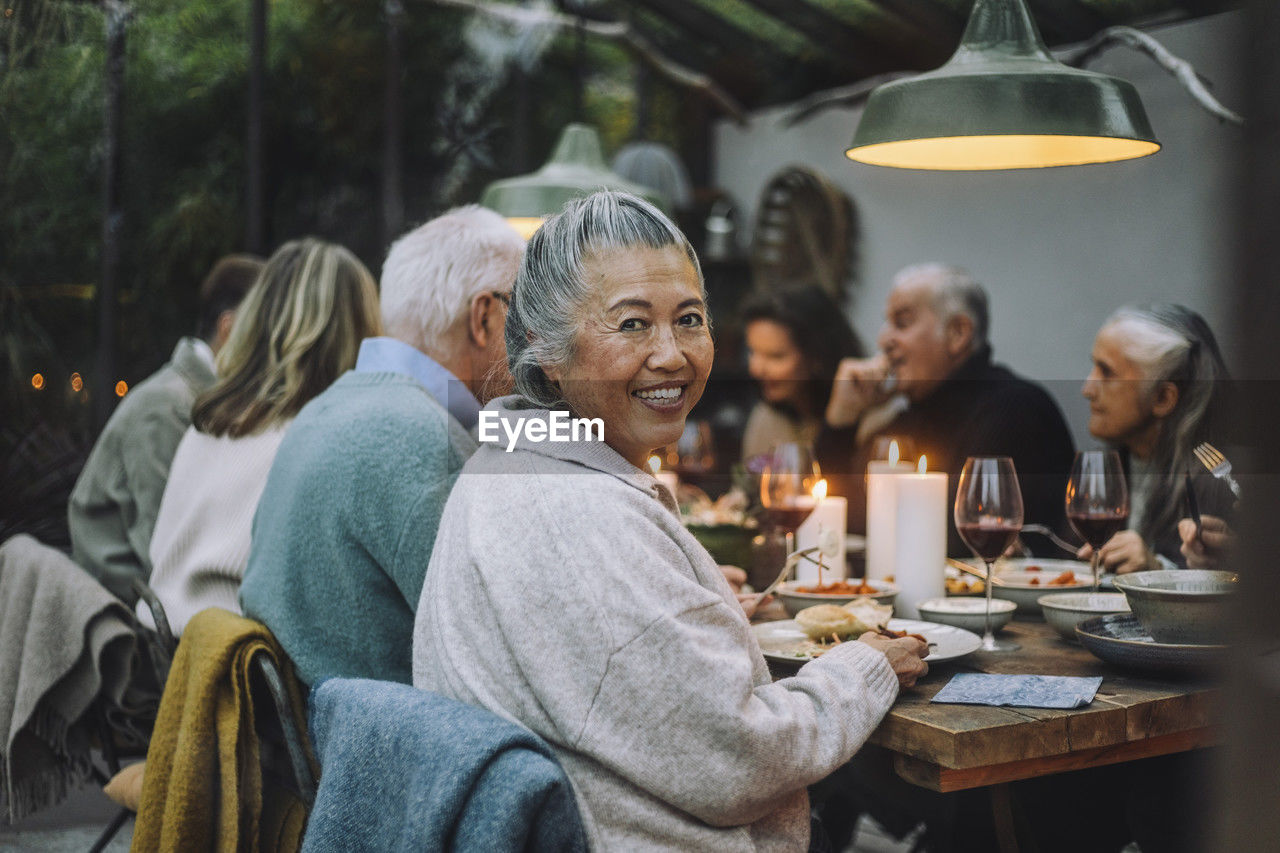 Smiling senior woman looking over shoulder while sitting with friends at dinner party