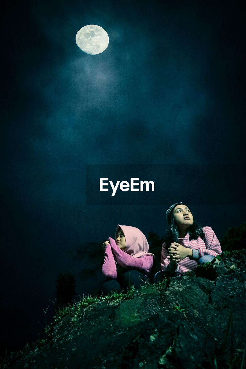 Low angle view of idyllic moon over friends sitting on rock at night