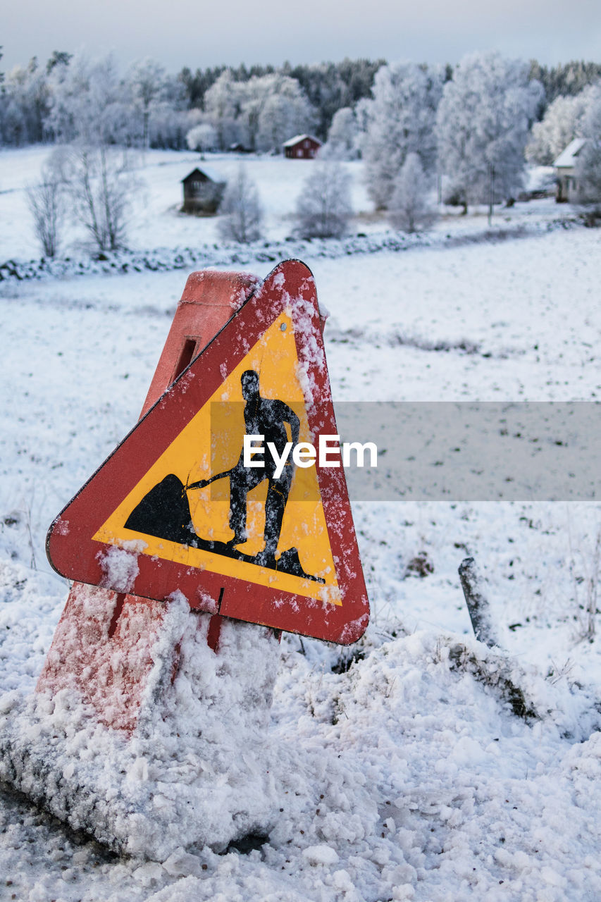 Construction sign covered in snow by a countryside road with winter landscape in the background