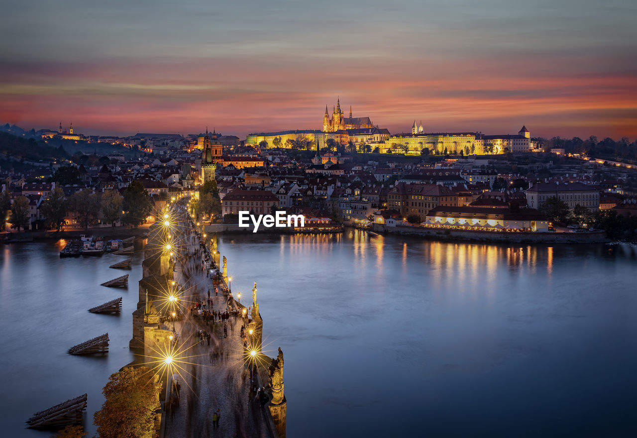 Illuminated cityscape by river against sky during sunset