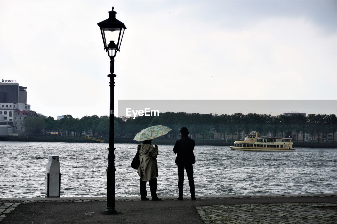 Rear view of man and woman standing on pier by river against sky
