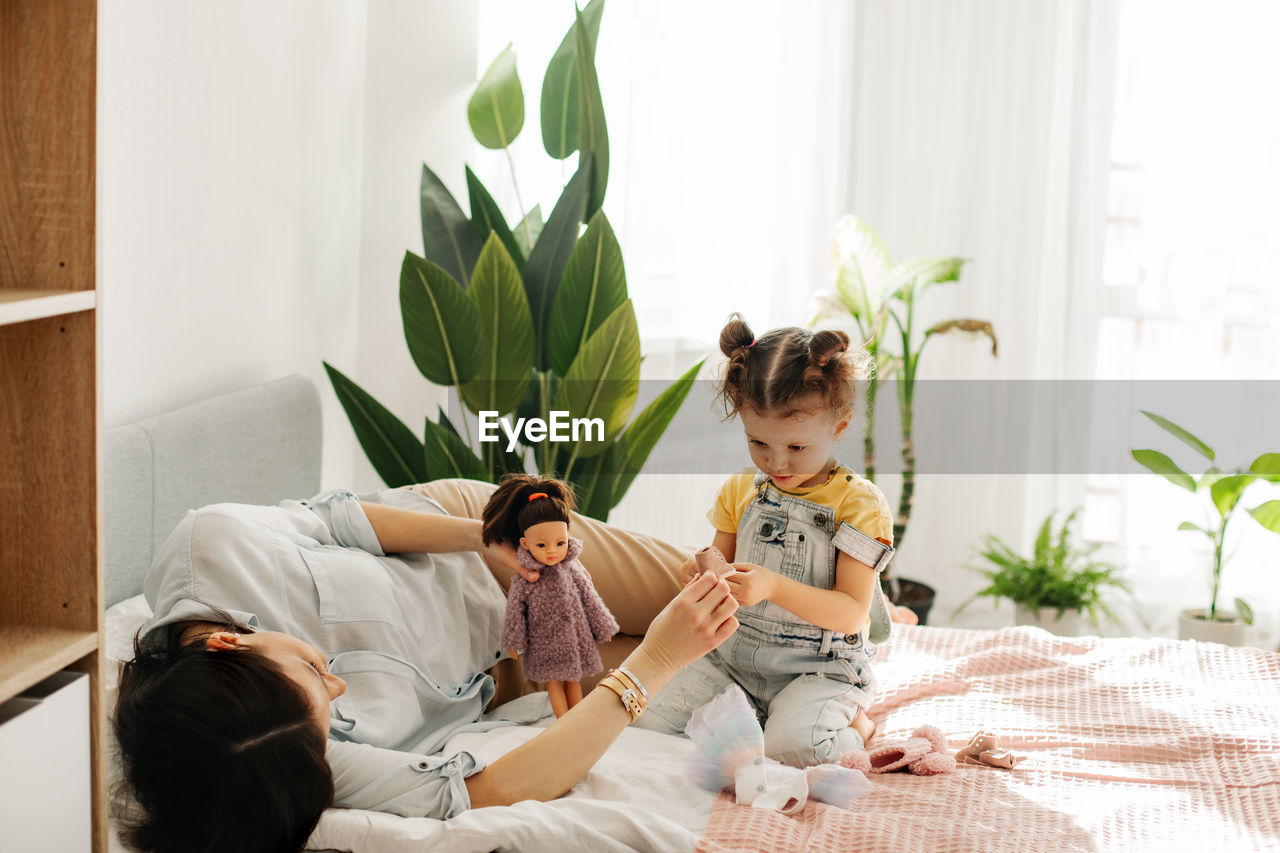 A charming little girl plays with dolls with her mom on the bed during the day. motherhood, caring.