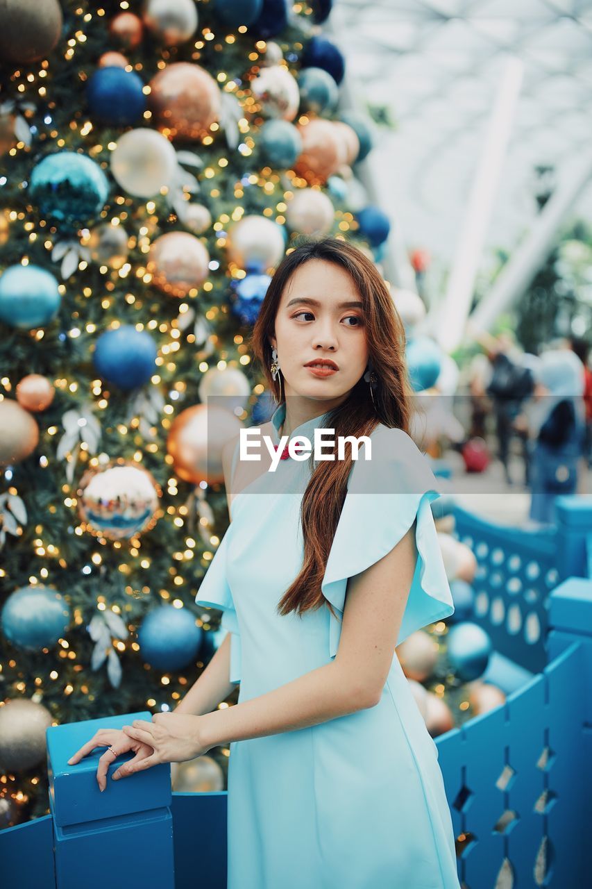 Smiling young woman looking away while standing by christmas tree outdoors