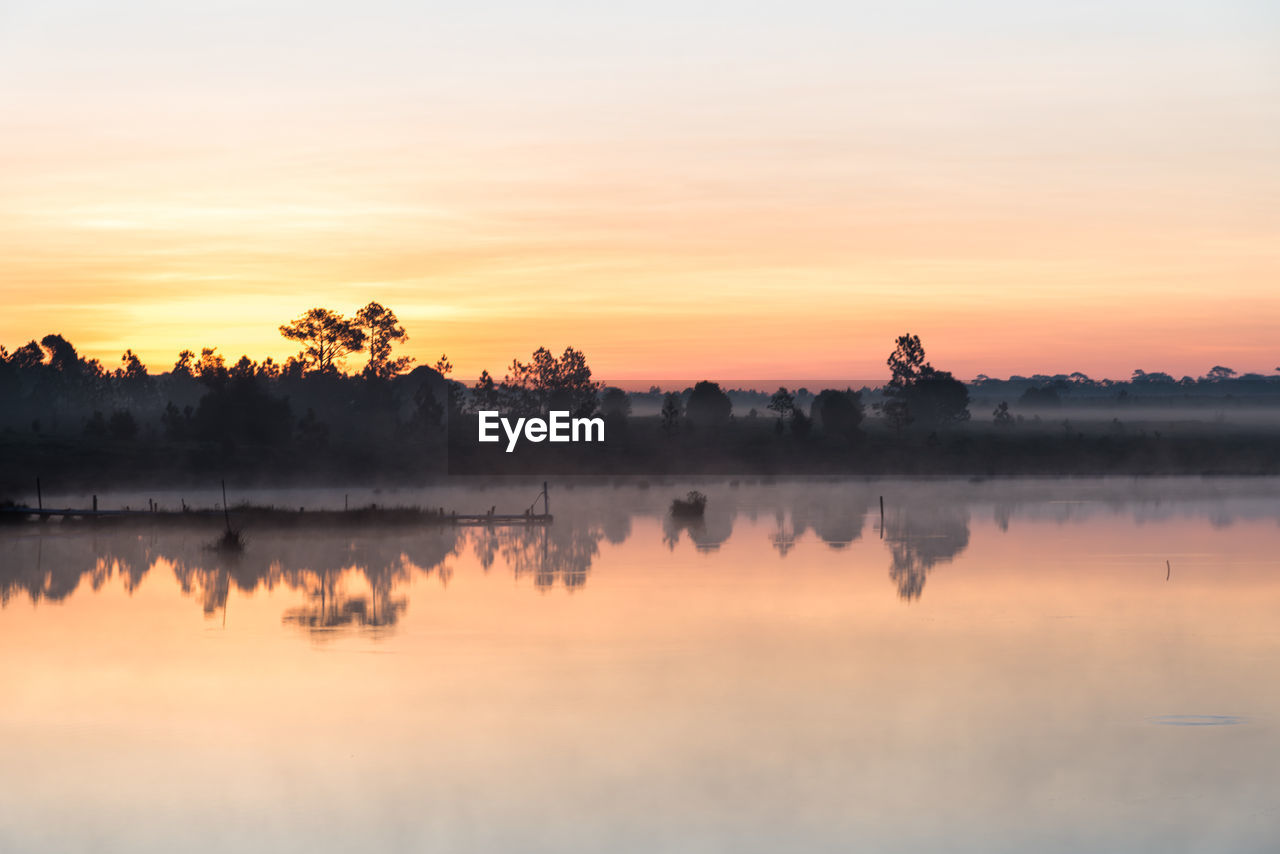 REFLECTION OF SILHOUETTE TREES IN LAKE AGAINST SKY DURING SUNSET