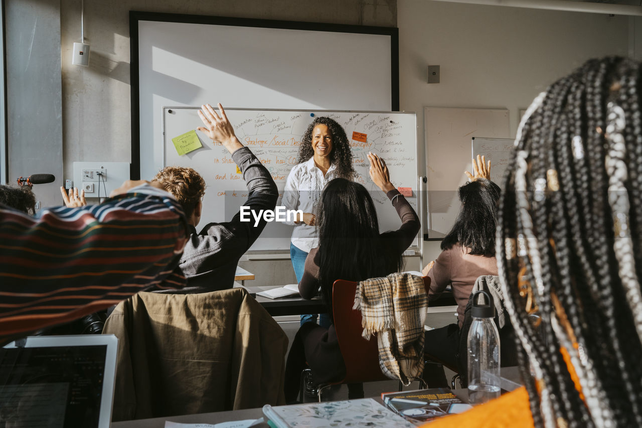 Smiling professor looking at university students sitting with hands raised in classroom