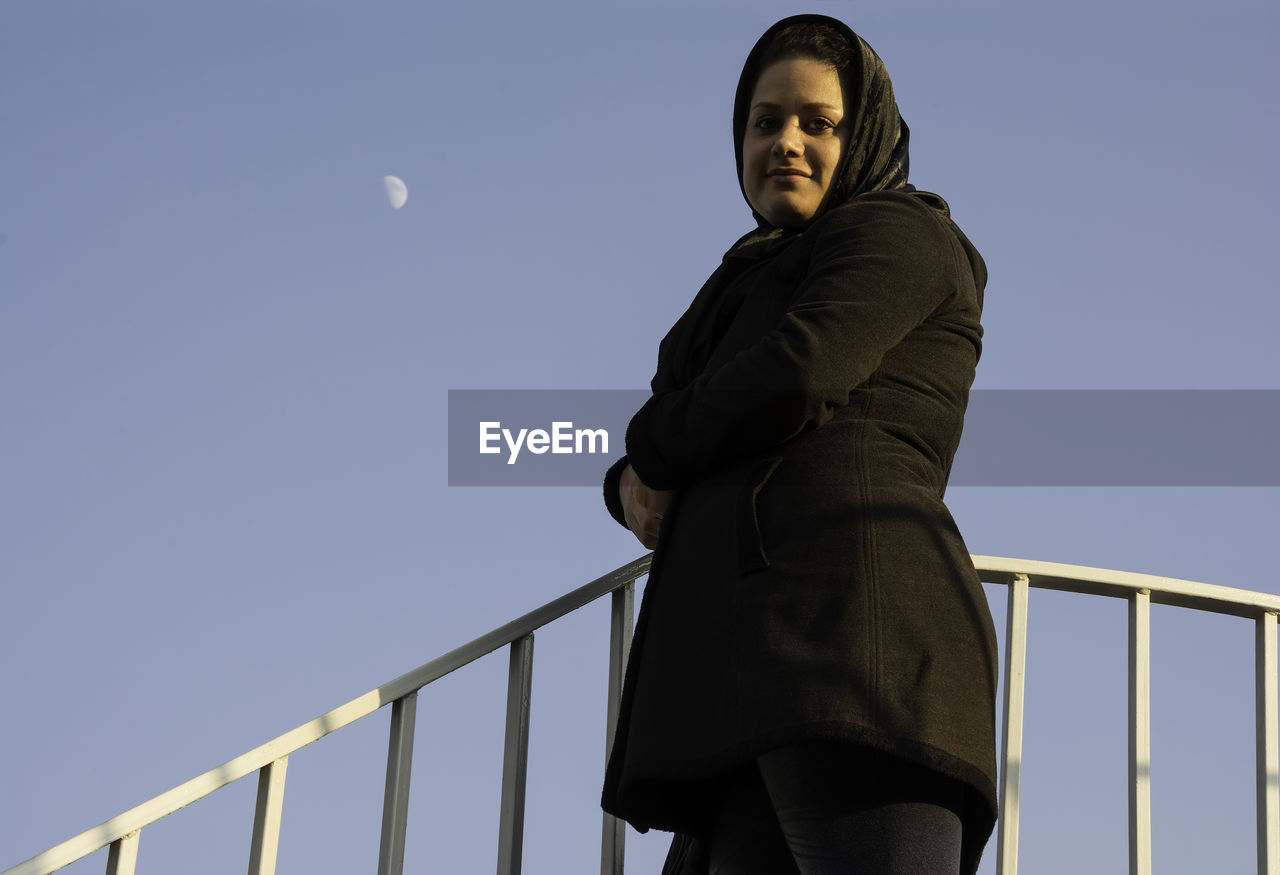 LOW ANGLE VIEW OF WOMAN STANDING ON RAILING AGAINST SKY