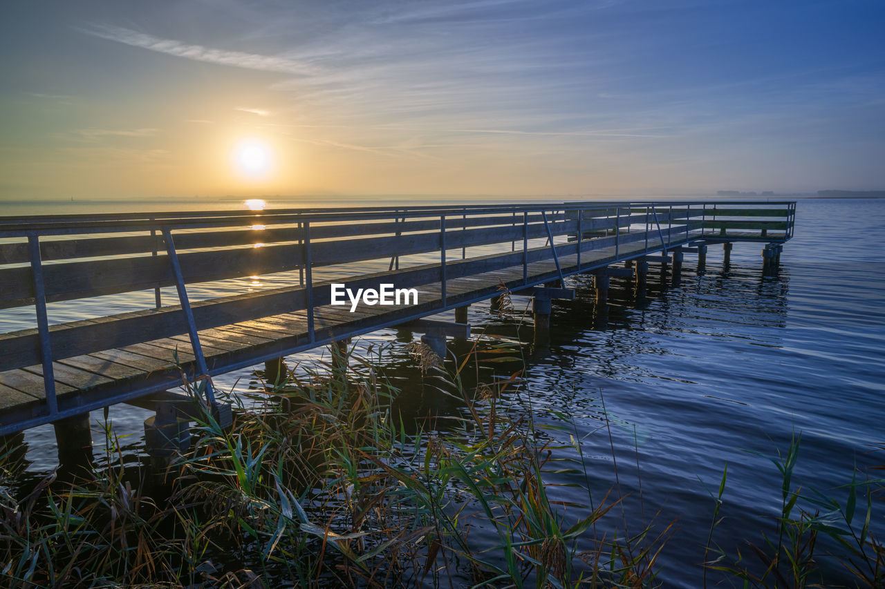 water, sky, reflection, horizon, sea, ocean, nature, beauty in nature, sunrise, scenics - nature, sunlight, tranquility, coast, shore, morning, tranquil scene, land, dawn, wave, no people, horizon over water, beach, sun, cloud, idyllic, outdoors, railing, wood, pier, non-urban scene, architecture, plant, environment, landscape, seascape, grass, built structure, travel destinations, blue