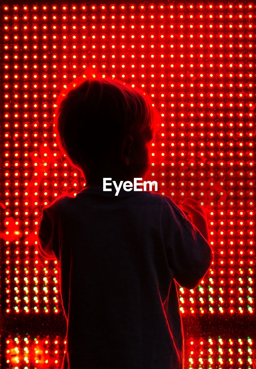 Rear view of boy standing in front of illuminated glass