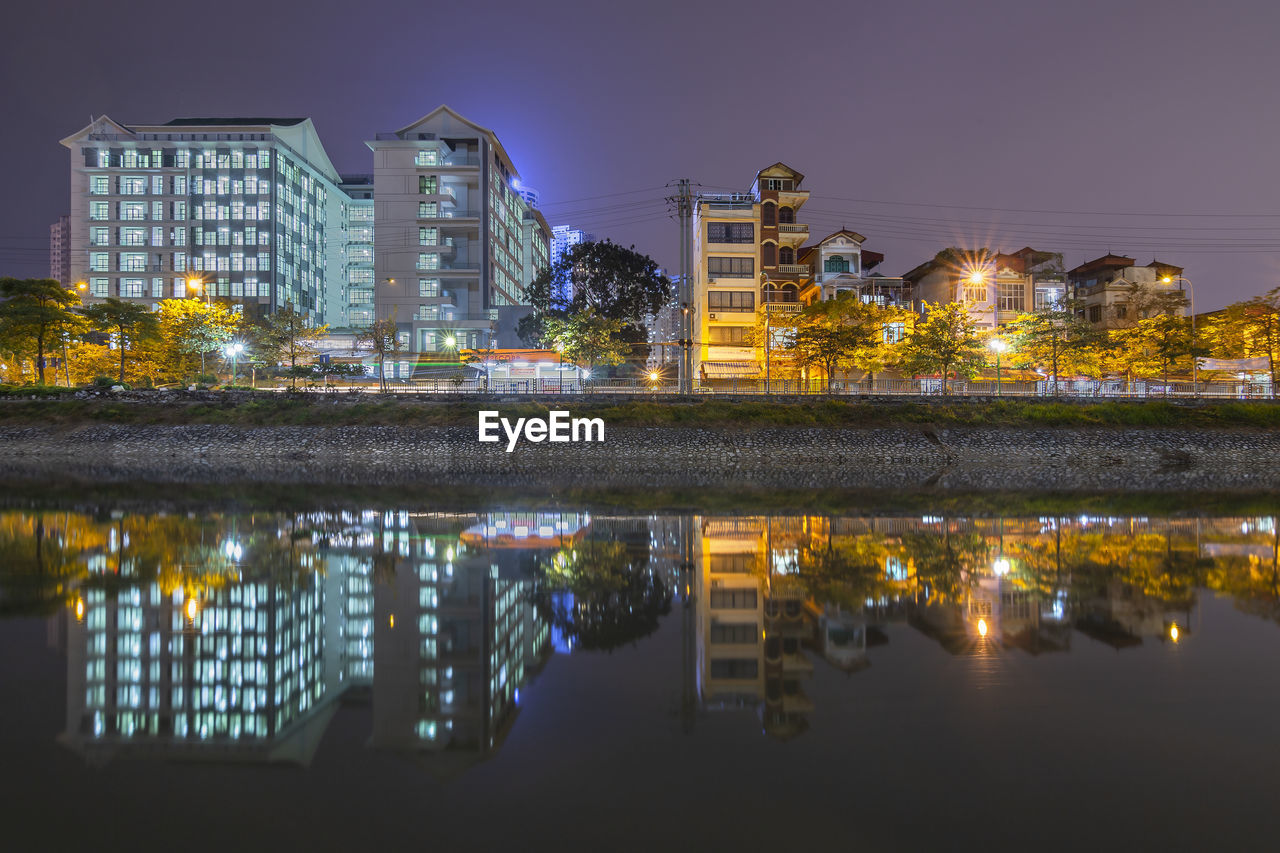 Illuminated buildings by lake against sky in city at night