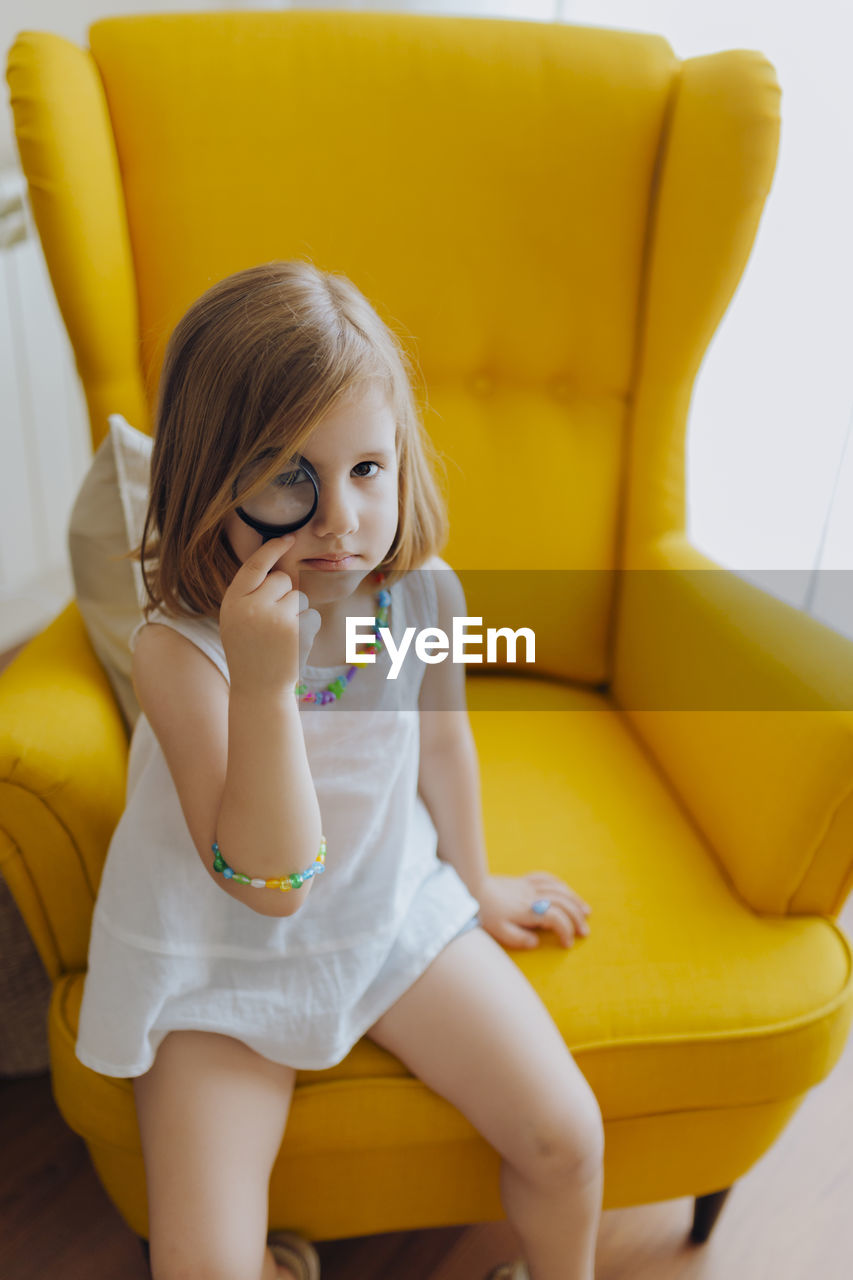 Blonde girl with a magnifying glass in her eye sitting on a yellow armchair