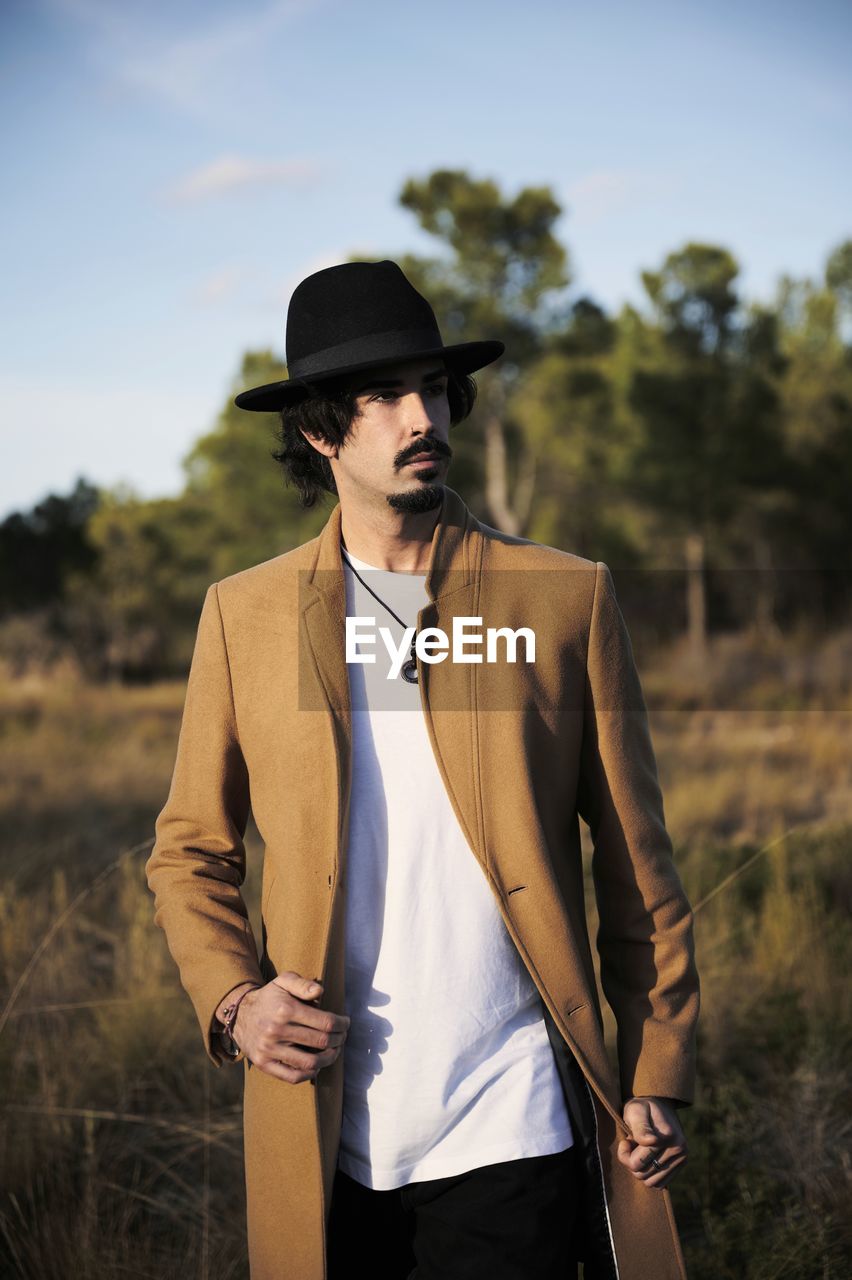 Young stylish hipster bearded male traveler with piercing wearing black hat looking away while standing on blurred countryside