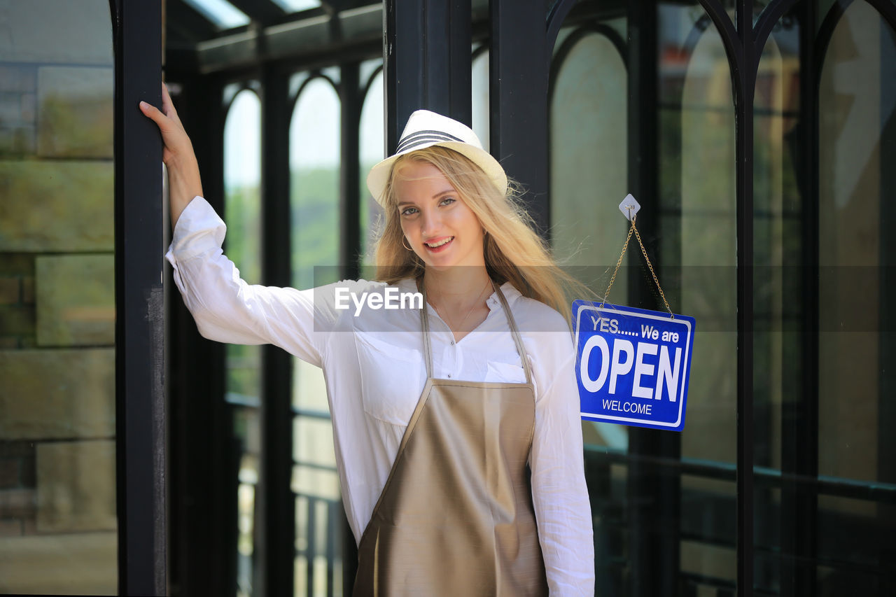 Portrait of young female owner of small business coffee shop standing at shop front with open sign.