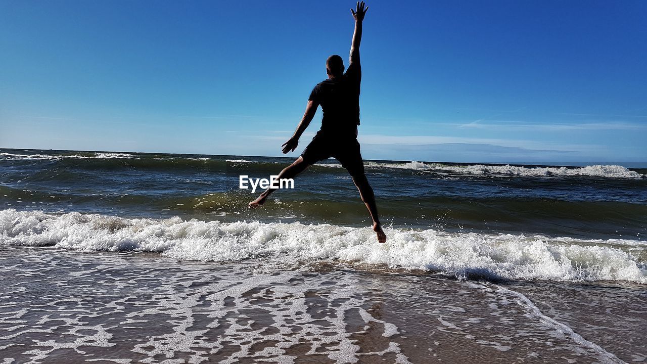 Rear view of man jumping on shore at beach against sky