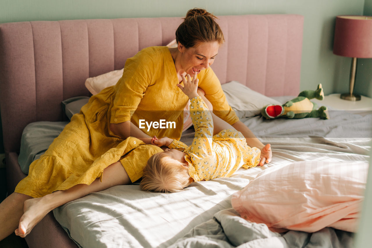 Mom tickles and hugs her little daughter in the morning on the bed in the apartment.