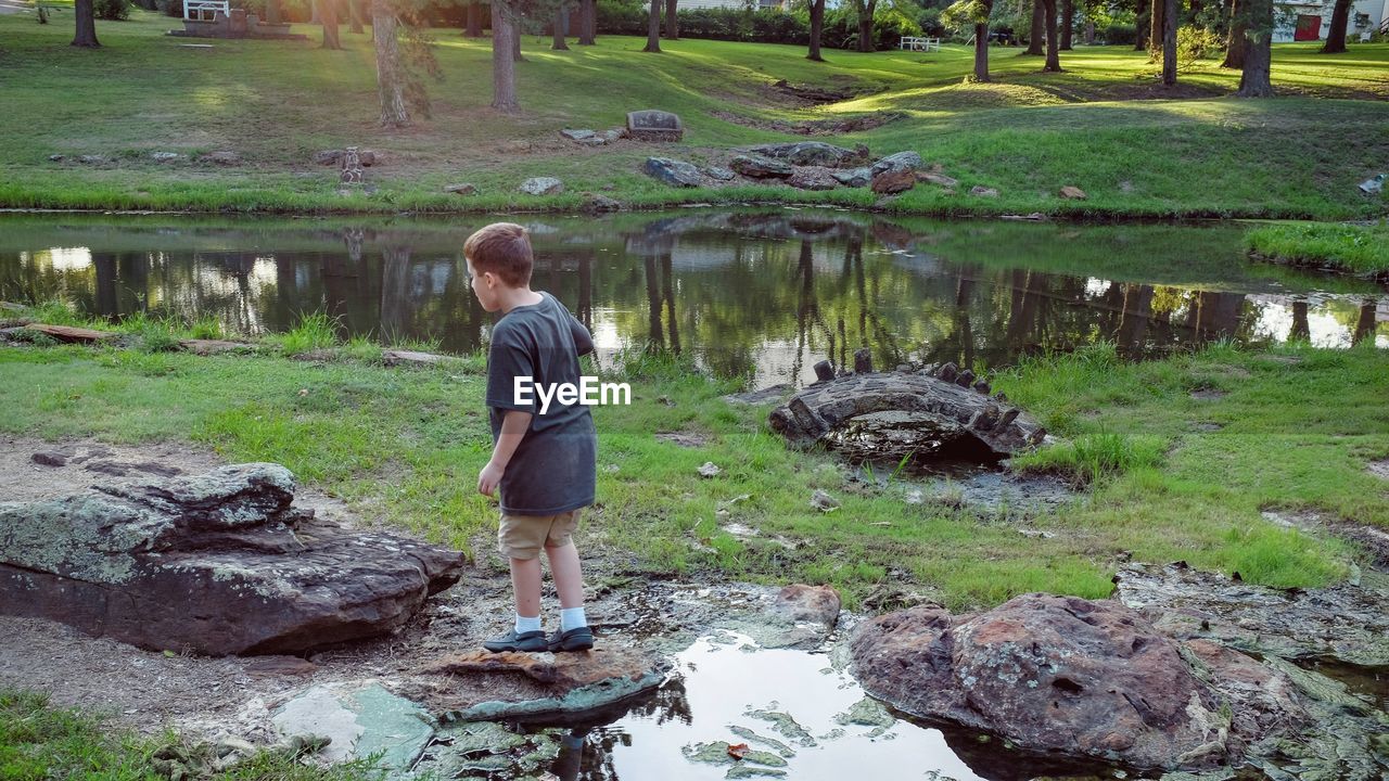 Full length of boy standing on rock by pond against trees in park