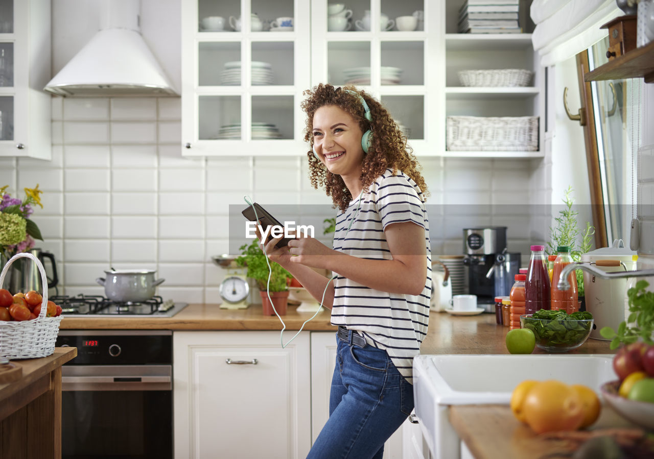 Side view of cheerful teenage girl listening music in kitchen at home