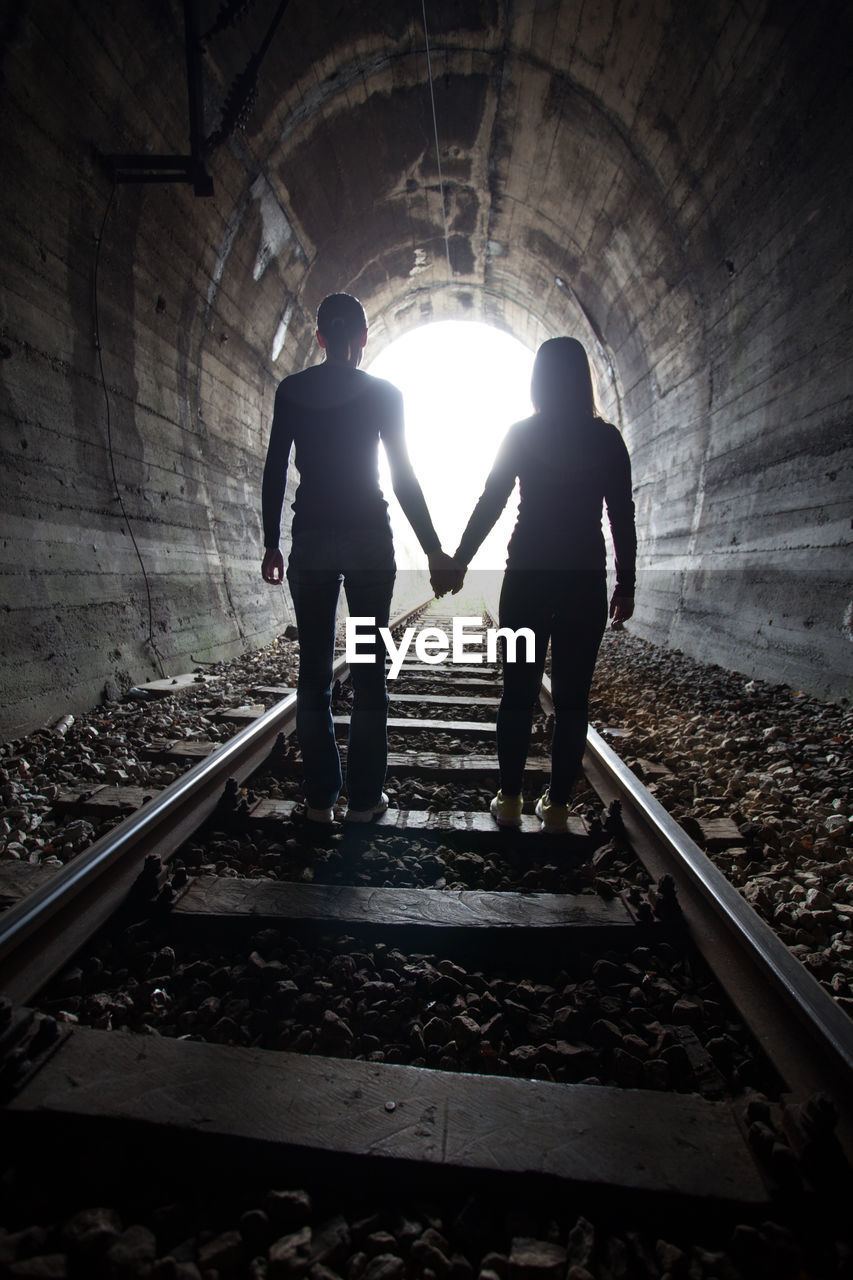 REAR VIEW OF SILHOUETTE MAN AND WOMAN STANDING ON RAILROAD TRACKS