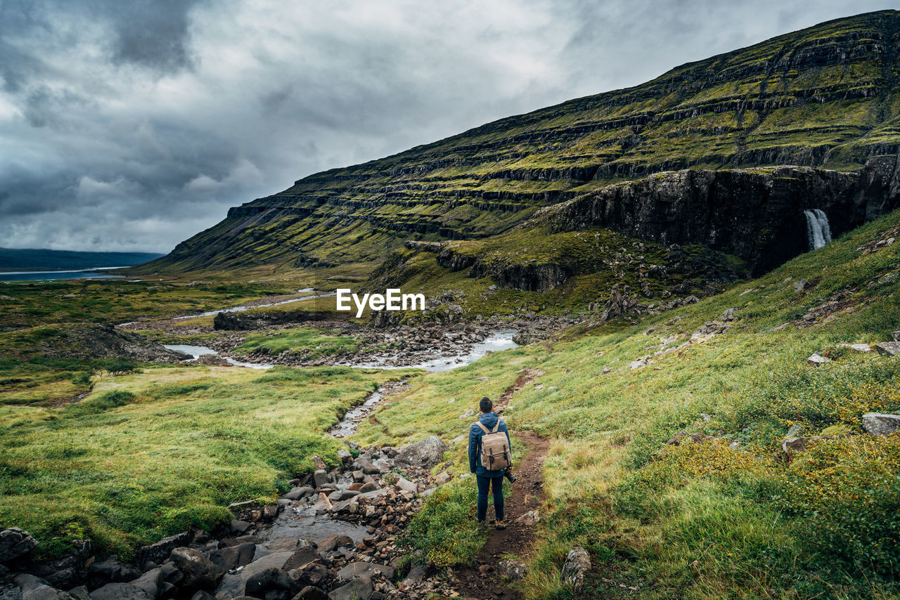 Full length rear view of man standing by mountains against cloudy sky