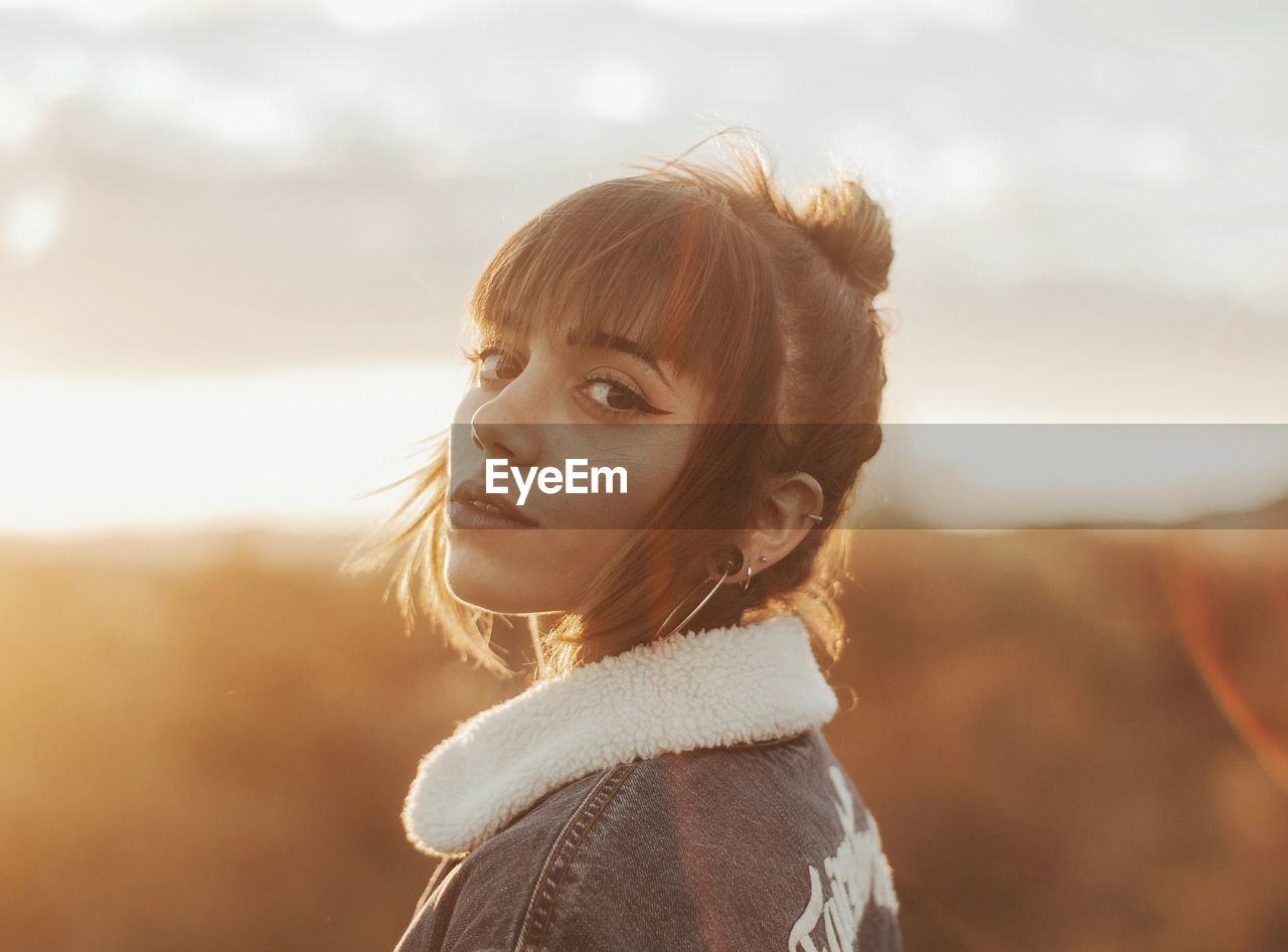 Close-up portrait of young woman standing against cloudy sky during sunset