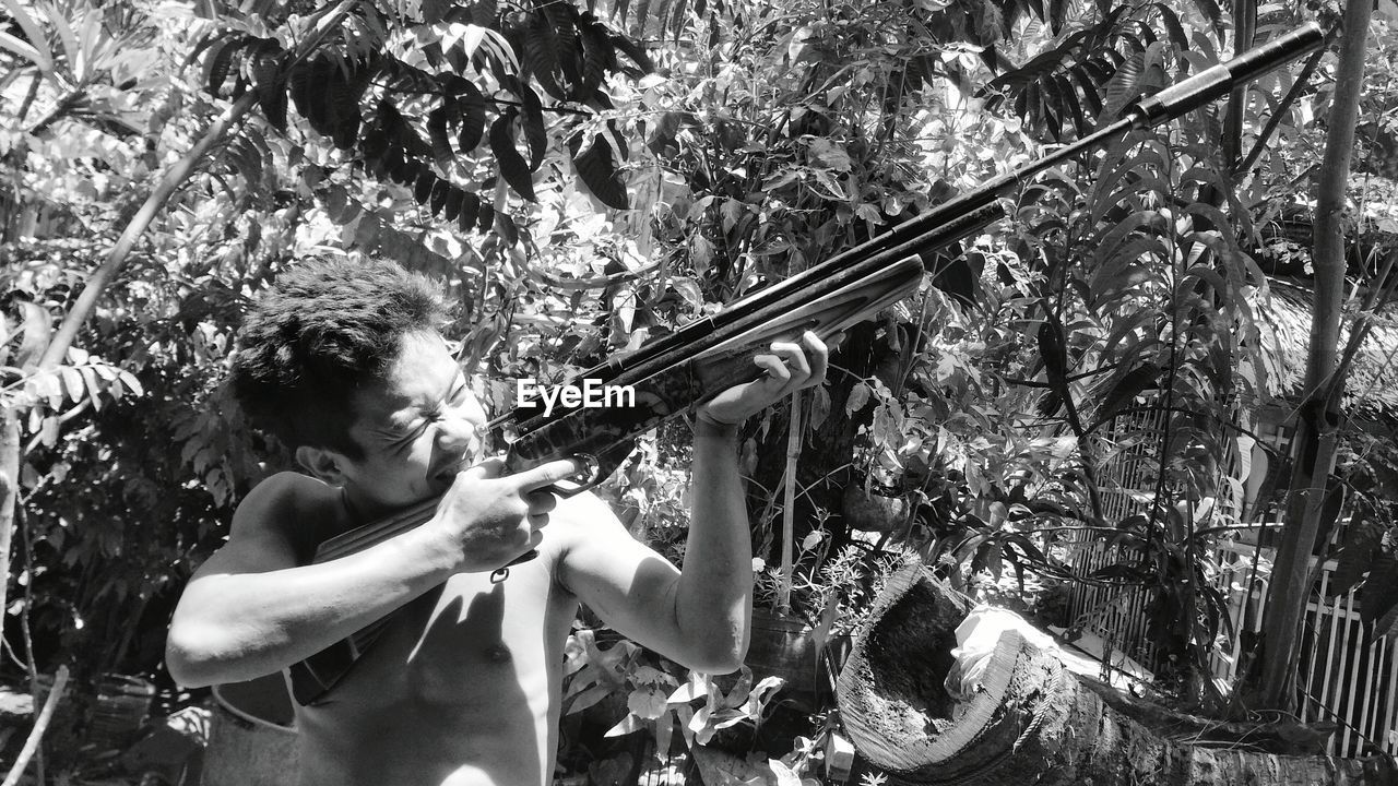 Young man aiming rifle while standing against plants