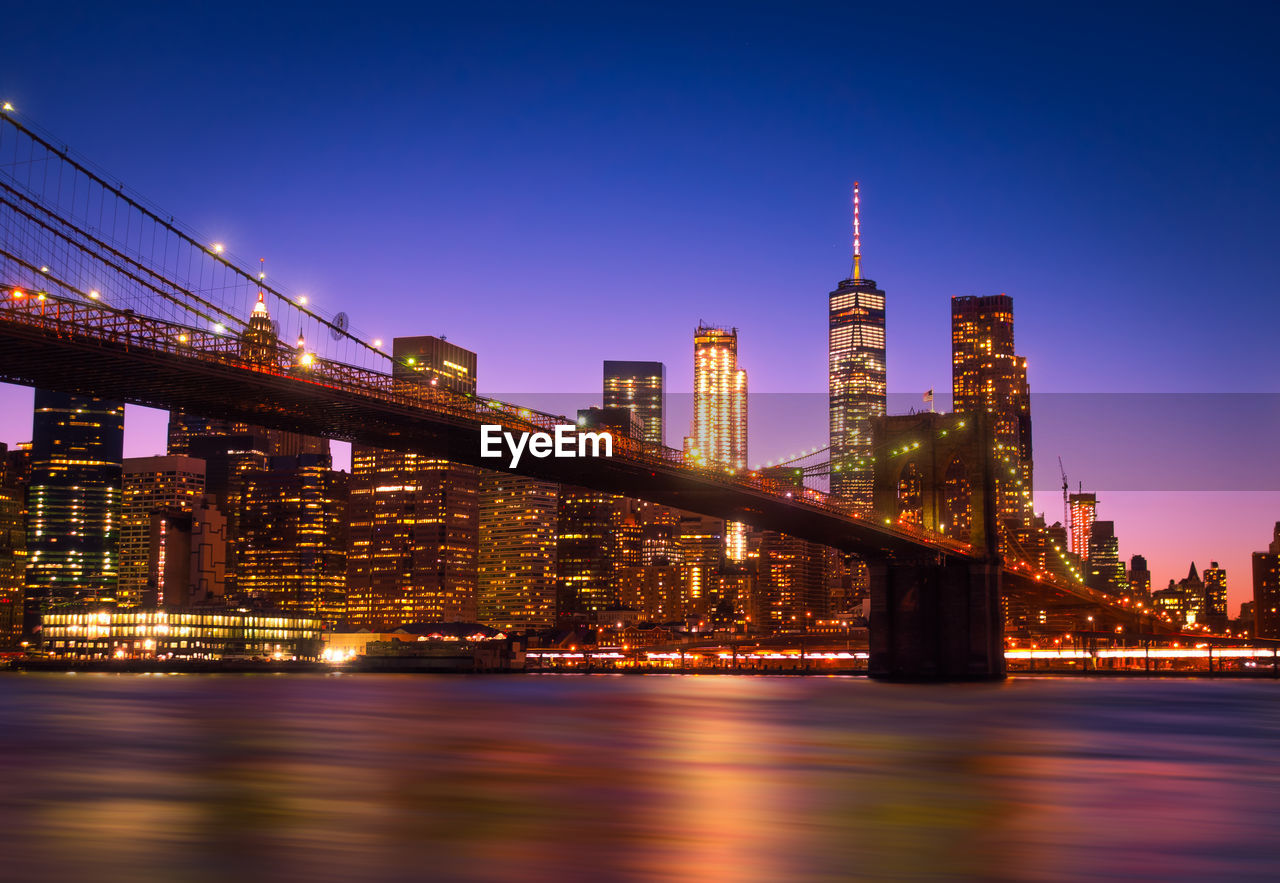 Illuminated bridge over river against buildings at night