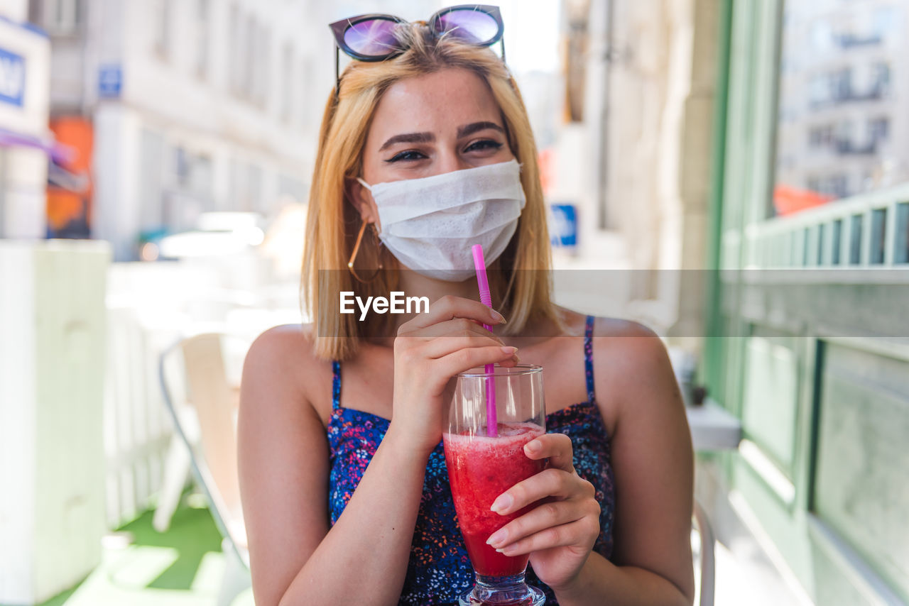 Portrait of young woman wearing mask drinking juice sitting at cafe