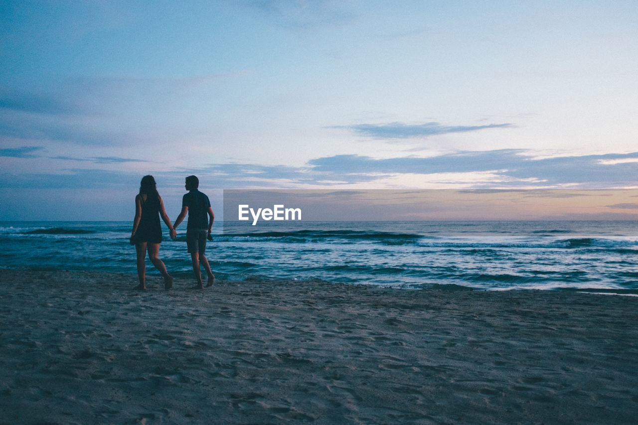 Silhouette couple walking on beach against sky during sunset