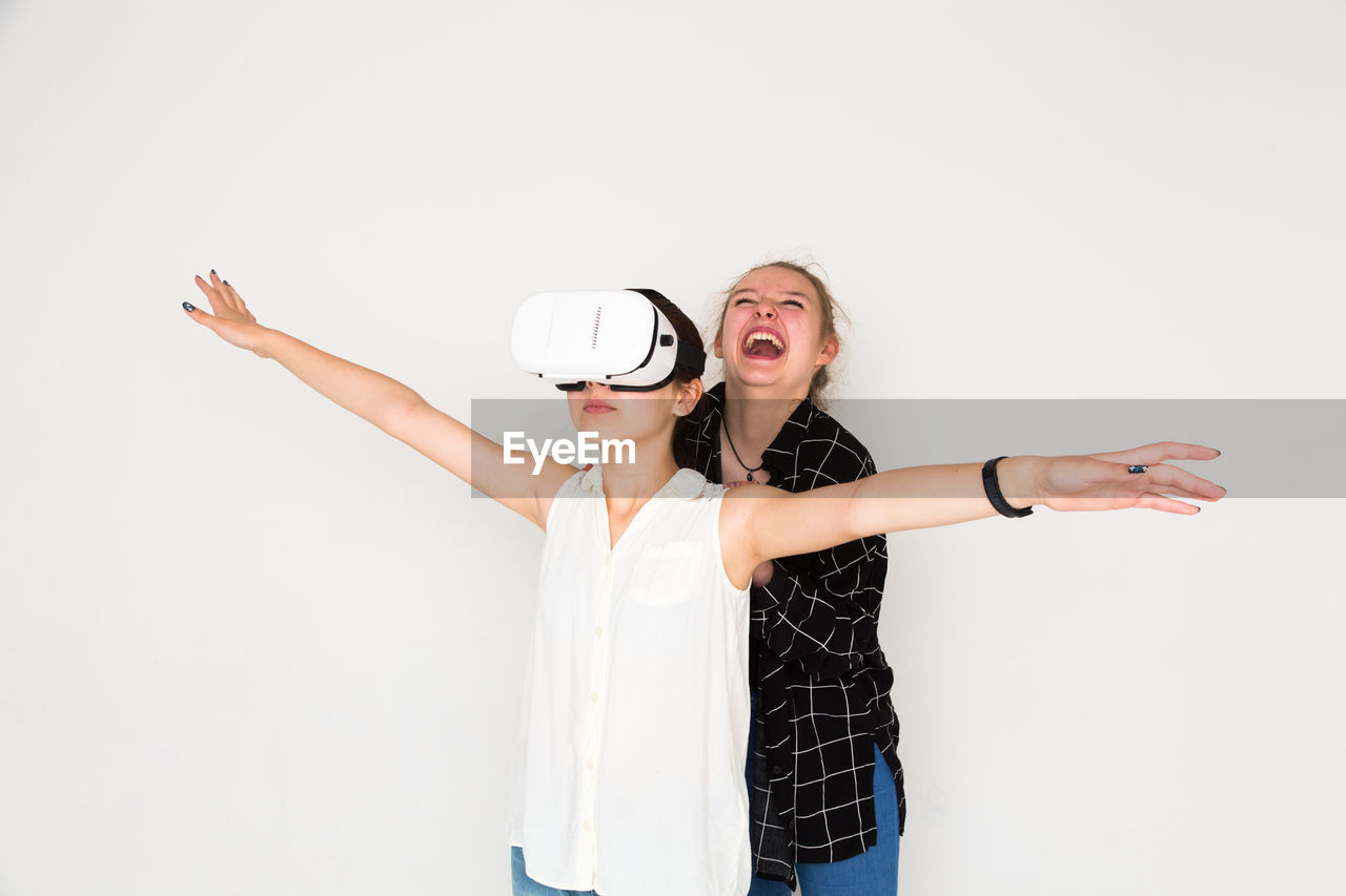 Cheerful woman standing by friend using virtual reality headset against white background