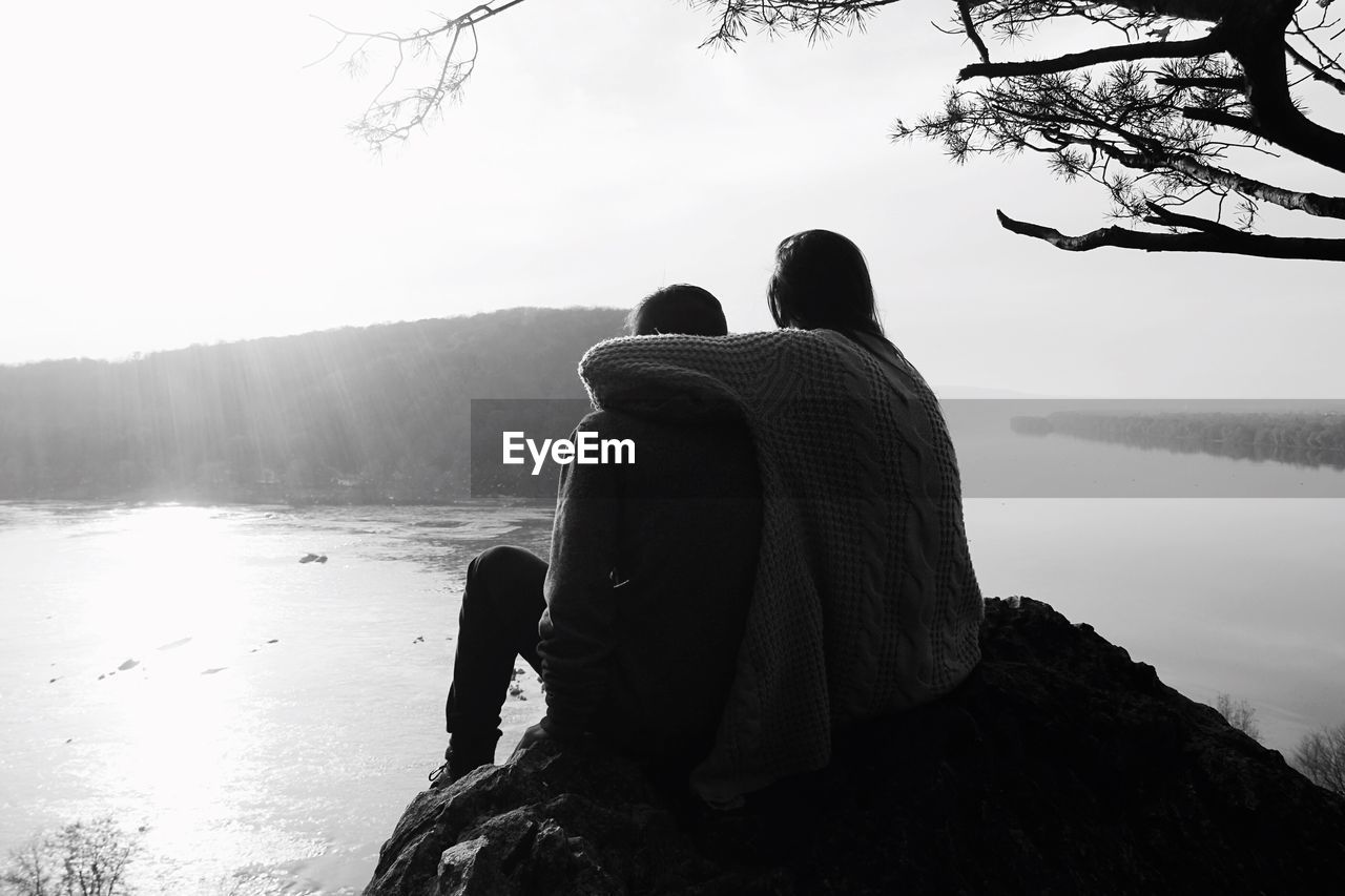 REAR VIEW OF COUPLE SITTING ON ROCK BY LAKE