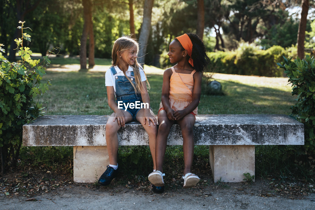 African american girl smiling and best friend sitting together on bench in green park in sunny day