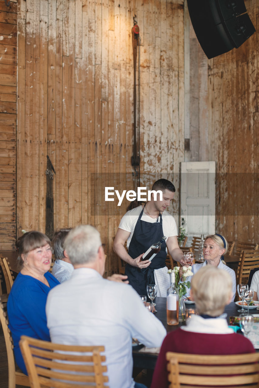 Young waiter serving wine to senior friends at table in restaurant