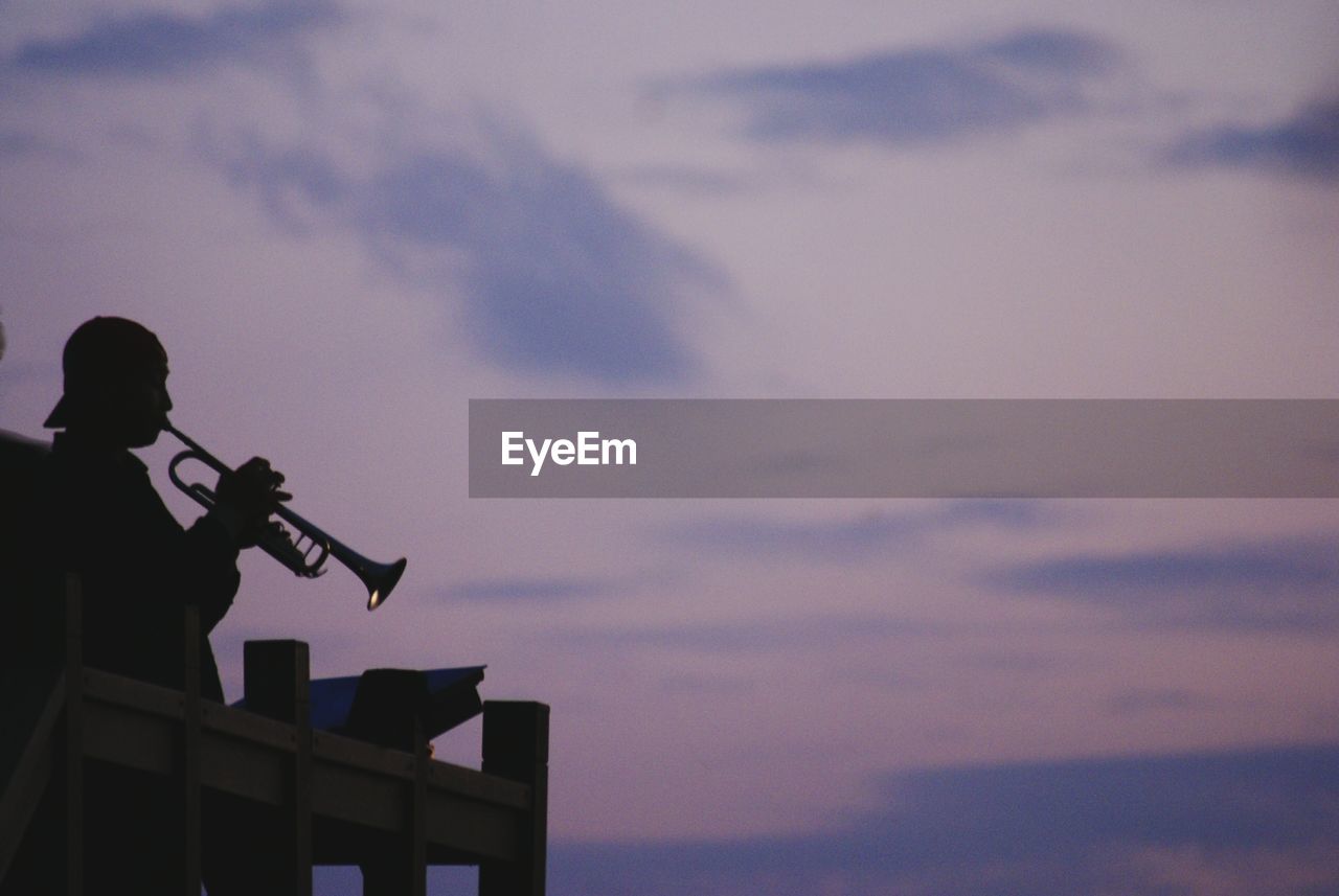 Silhouette man playing trumpet at observation point against cloudy sky during sunset