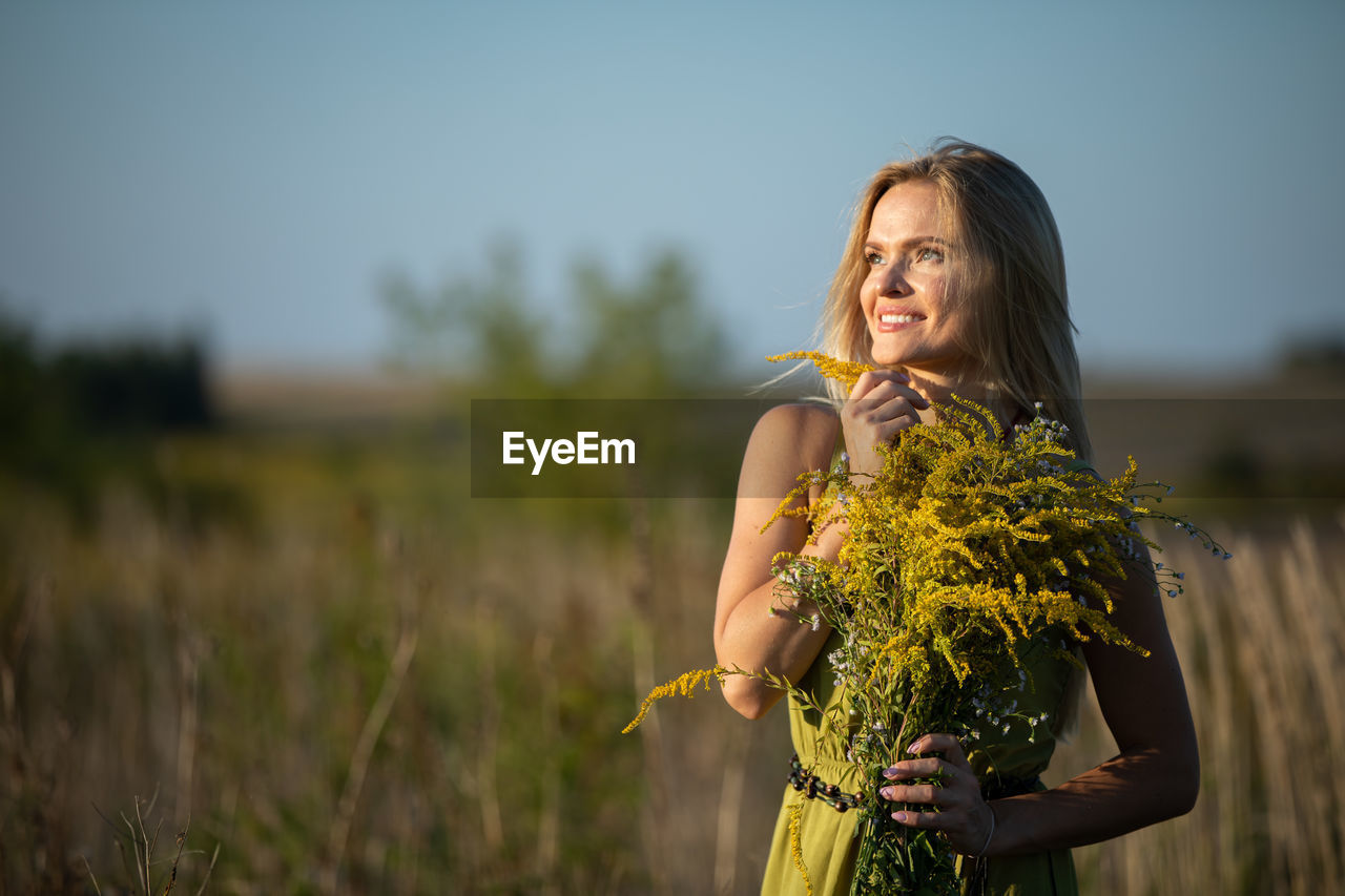 yellow, adult, women, plant, one person, nature, sky, flower, smiling, rural scene, young adult, flowering plant, landscape, happiness, emotion, long hair, hairstyle, dress, beauty in nature, female, clothing, field, land, environment, outdoors, spring, standing, fashion, portrait, summer, grass, autumn, lifestyles, agriculture, freshness, copy space, crop, focus on foreground, holding, three quarter length, food and drink, food, blond hair, sunlight, person, cheerful, photo shoot, portrait photography, day, flower arrangement, wellbeing, waist up, looking, sunset, positive emotion, bouquet