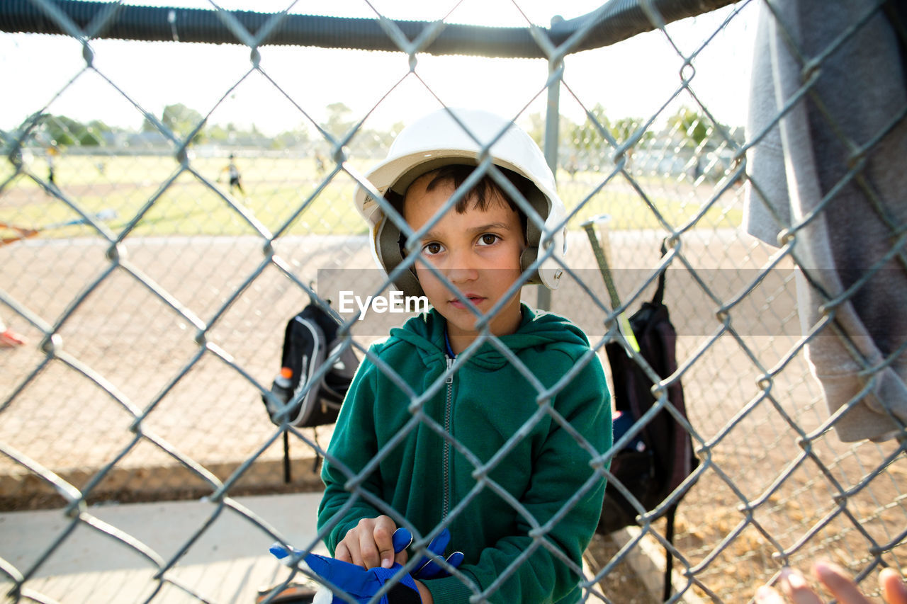 Portrait of boy wearing baseball helmet while sitting in dugout seen through chainlink fence