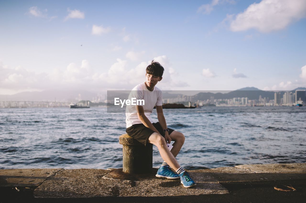 Young man sitting on bollard by sea against sky