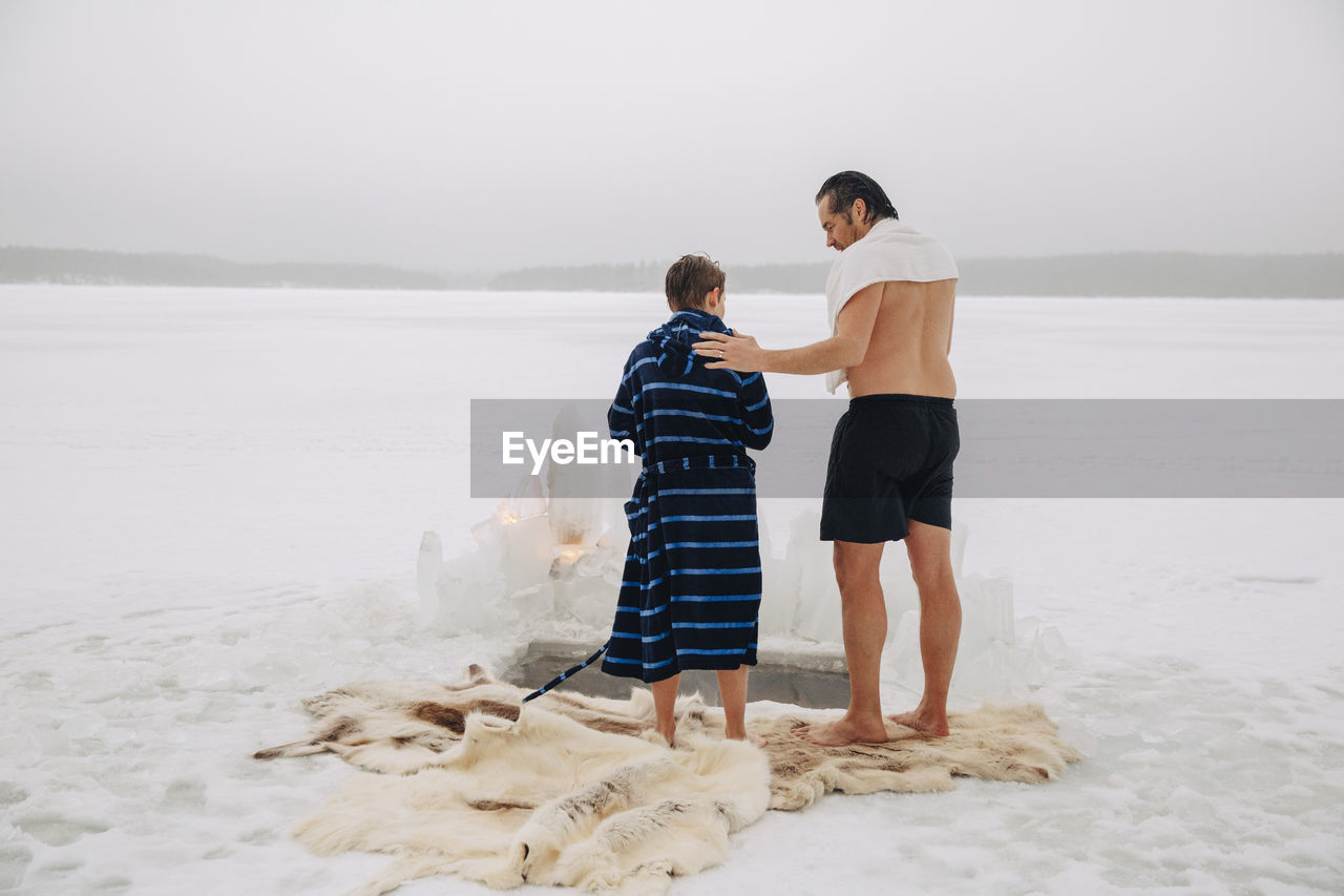 Rear view of shirtless man talking with son wearing bathrobe while standing on animal skin at frozen lake