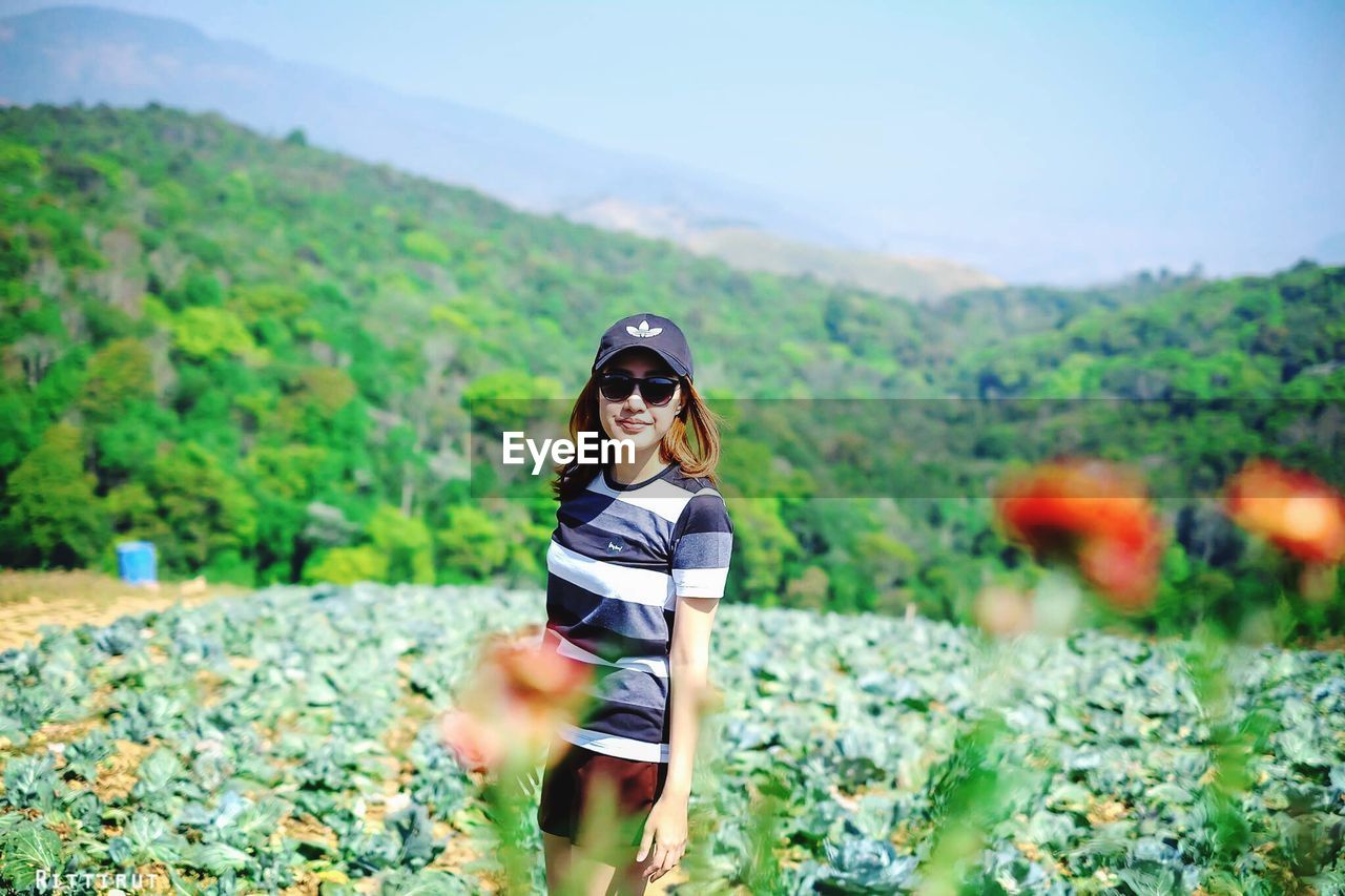 PORTRAIT OF YOUNG WOMAN IN SUNGLASSES STANDING ON LANDSCAPE