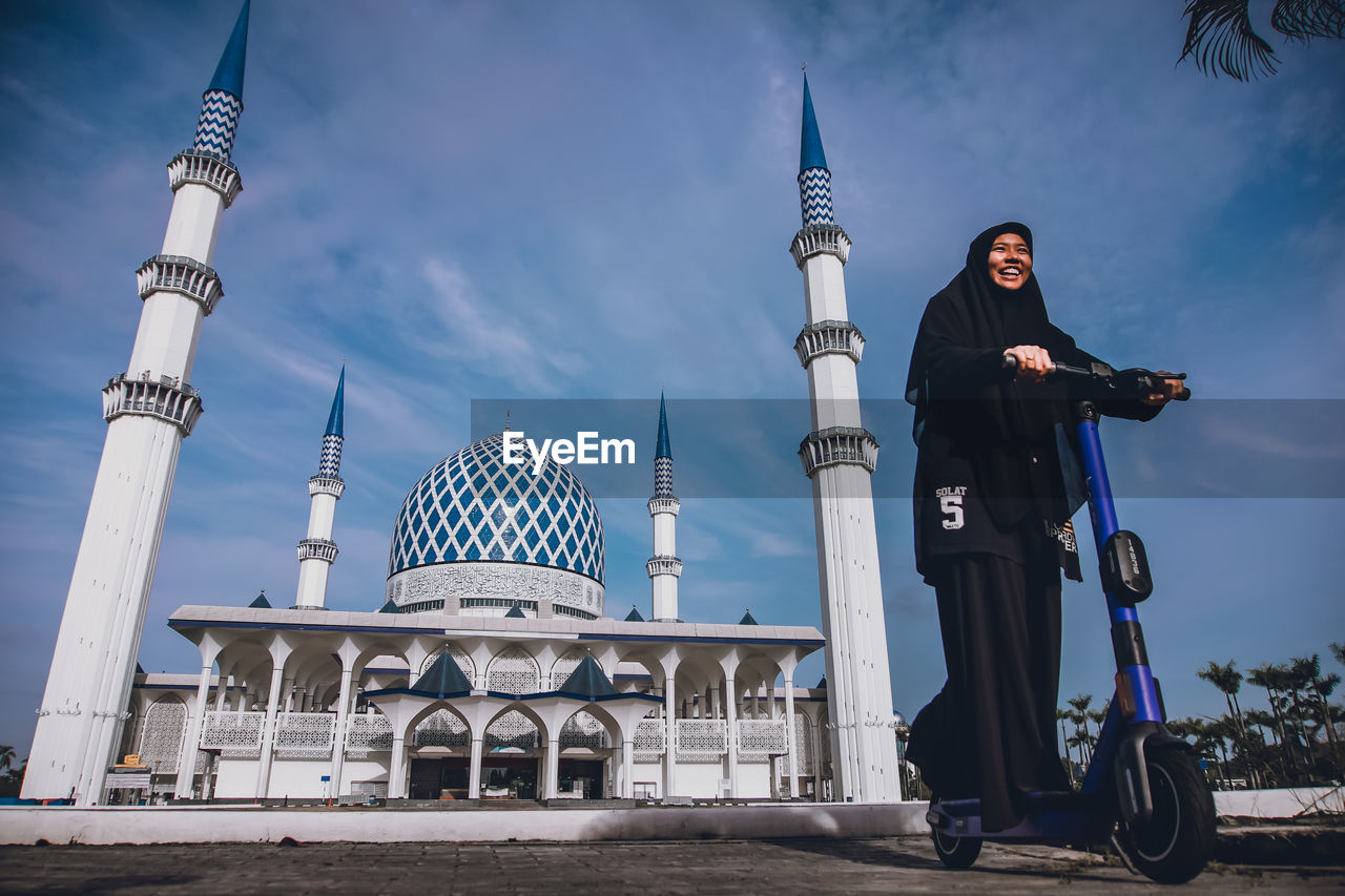 Low angle view of smiling woman riding push scooter against mosque