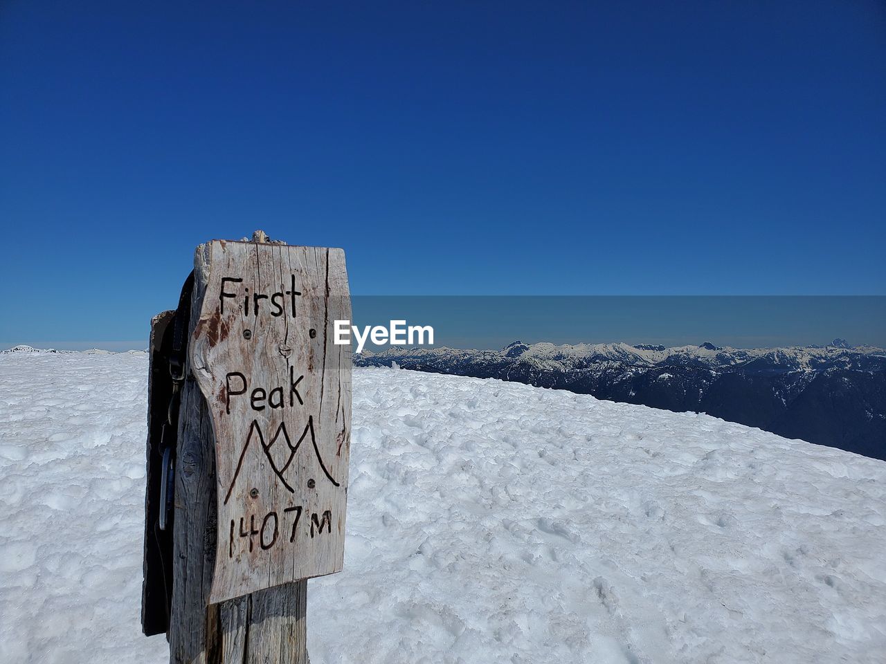 Mountain peak sign with text on snow covered landscape against blue sky