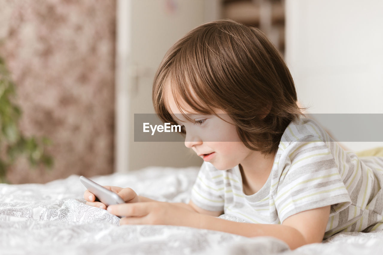 A cute boy, toddler, lies on the bed in the bedroom and plays with the phone, smartphone. 