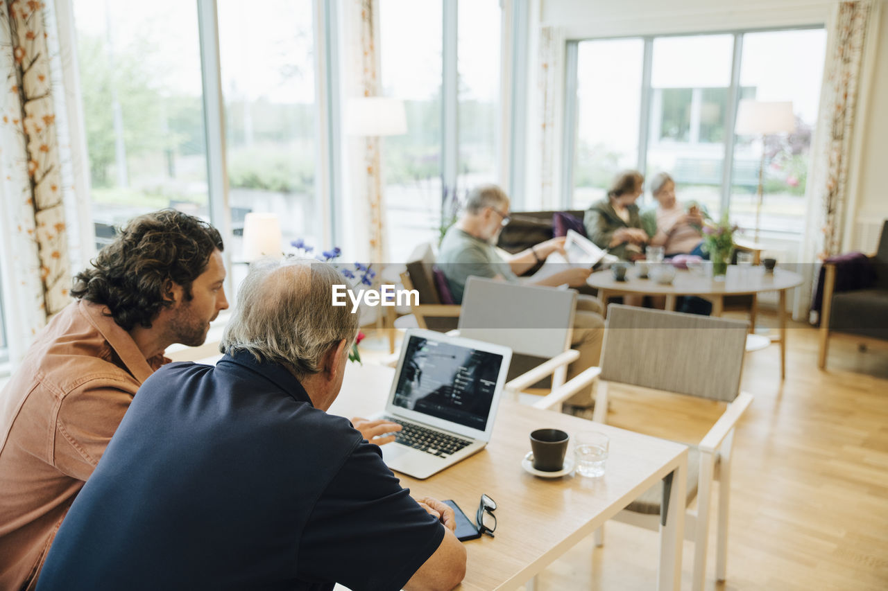 Man assisting elderly grandfather using laptop at dining table in retirement home