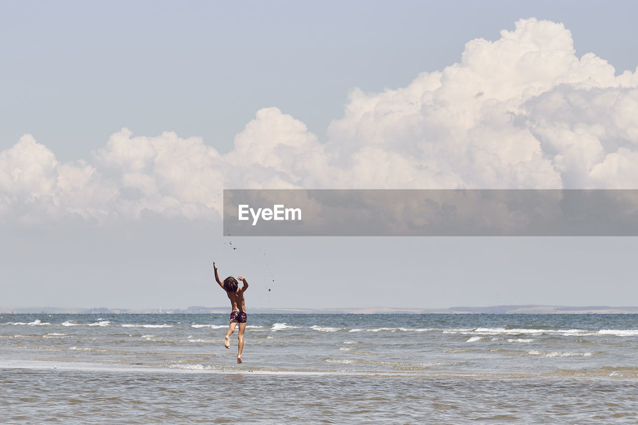 Rear view of shirtless boy jumping in sea against cloudy sky
