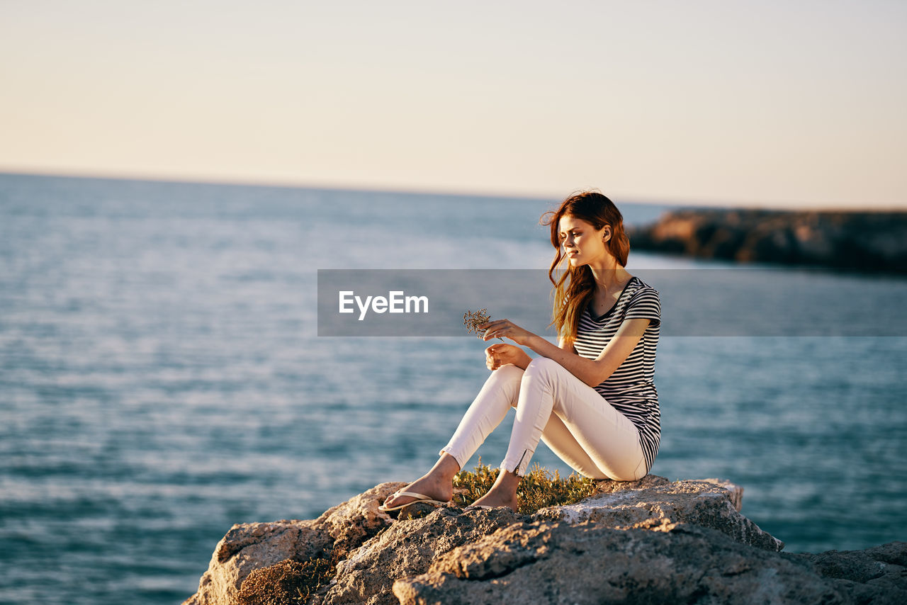 WOMAN SITTING ON ROCK AGAINST SEA