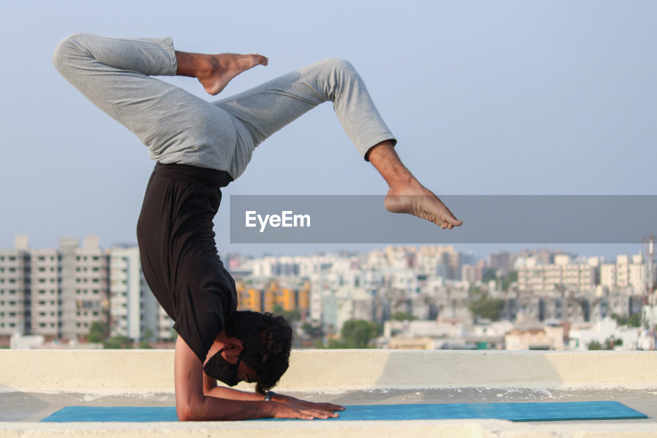 Side view of man doing yoga on highrise building terrace against sky