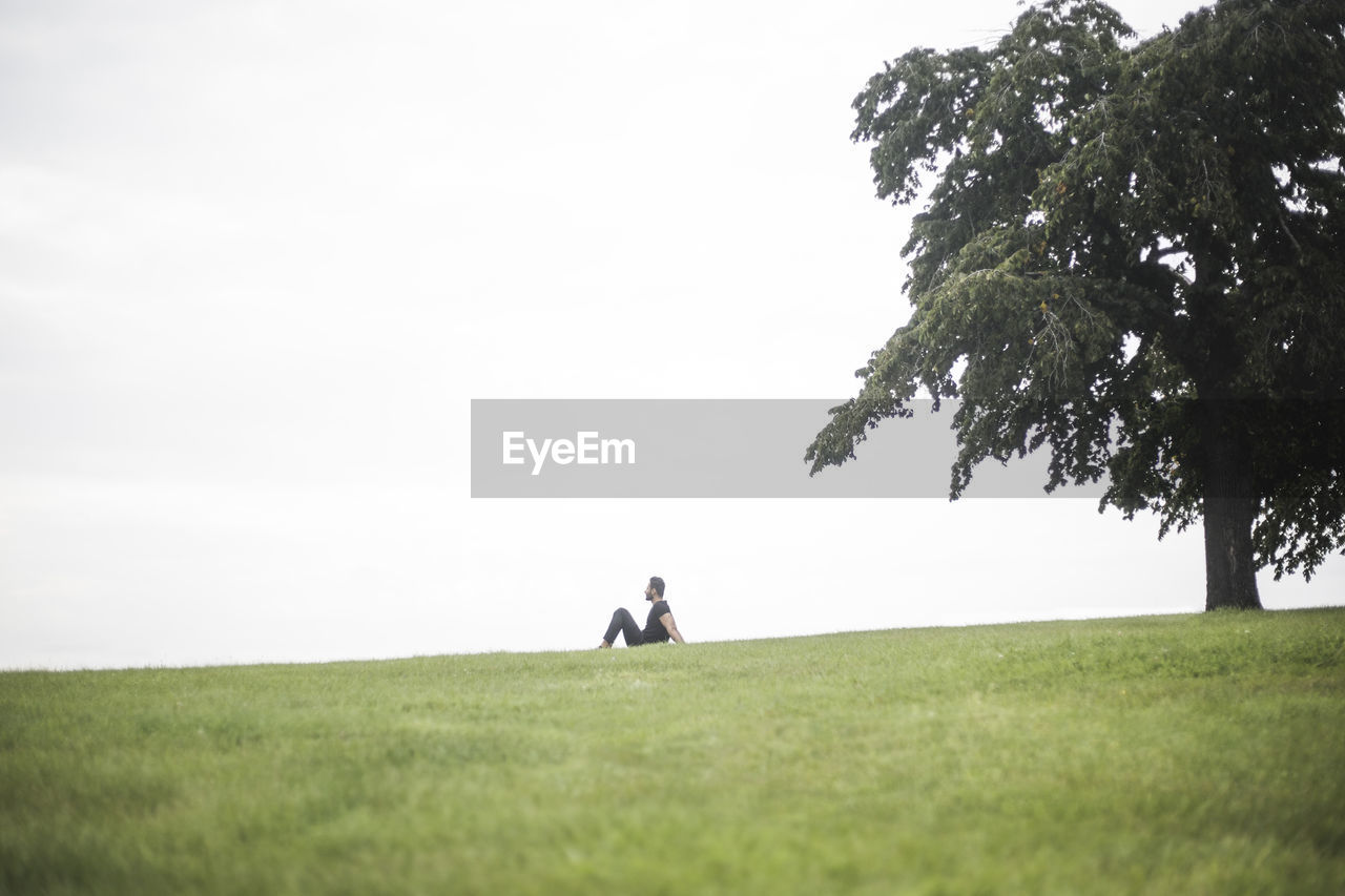 Mid distance view of man sitting on grassy field against sky