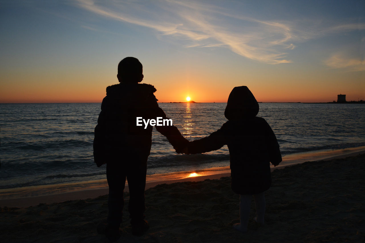 Silhouette siblings holding hands while standing at beach during sunset
