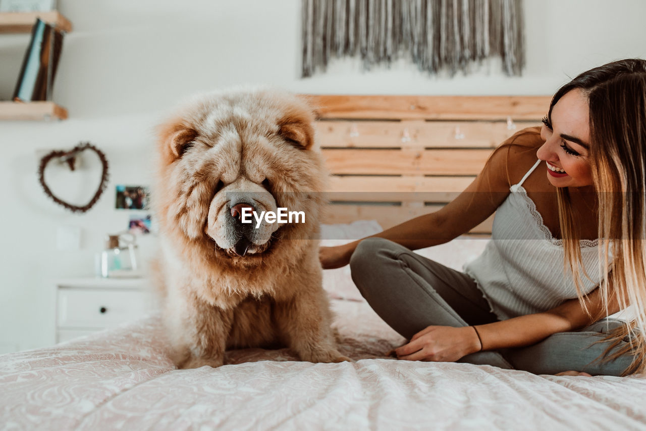 Smiling young woman sitting with dog on bed at home